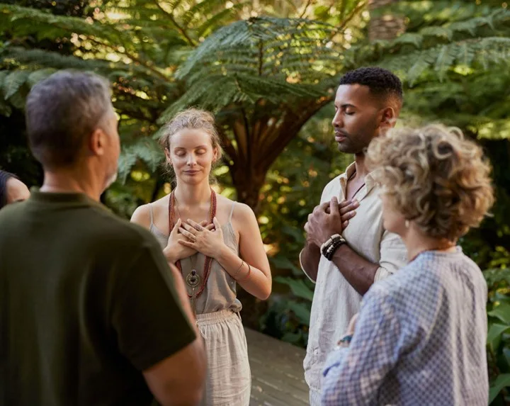 Mixed-raced group of adults standing peacefully in a circle with hands on their hearts and eyes closed during a wellness retreat in a forest setting.
