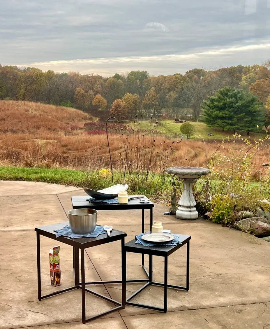 three small tables on a stone patio, each with candles and cloth on top. the patio overlooks a meadow with green, red, and brown foliage